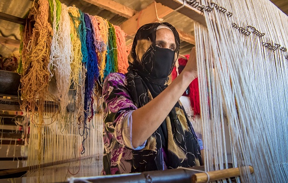 모로코의 노년 여성이 색색깔의 실을 배경으로 하여 수공으로 직물을 짜고 있는 사진